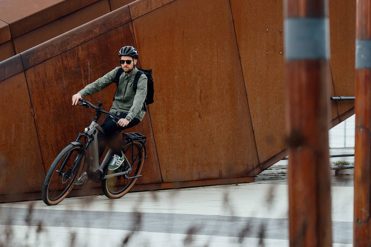 Man with helmet, sunglasses and rucksack riding a company bike in front of a rust-red wall