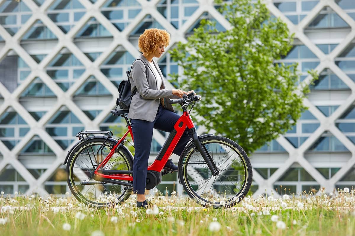 Frau steht mit roten Gazelle E-Bike auf Radweg dahinter modernes Gebäude und Baum