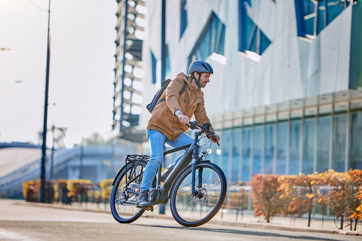 Mann mit Helm und Rucksack fährt Gazelle E-Bike entlang von modernem Gebäude