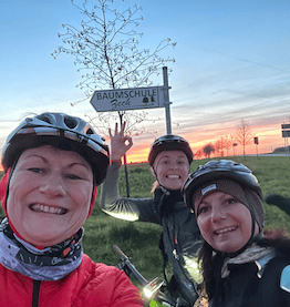 three women with helmets riding company bikes in front of sunset smiling towards camera