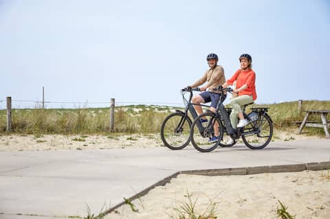 Paar auf Diensträdern auf Holzsteg im Hintergrund Dünenlandschaft