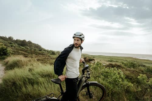 Mann mit Helm steht mit Dienstrad in Dünen und schaut zurück