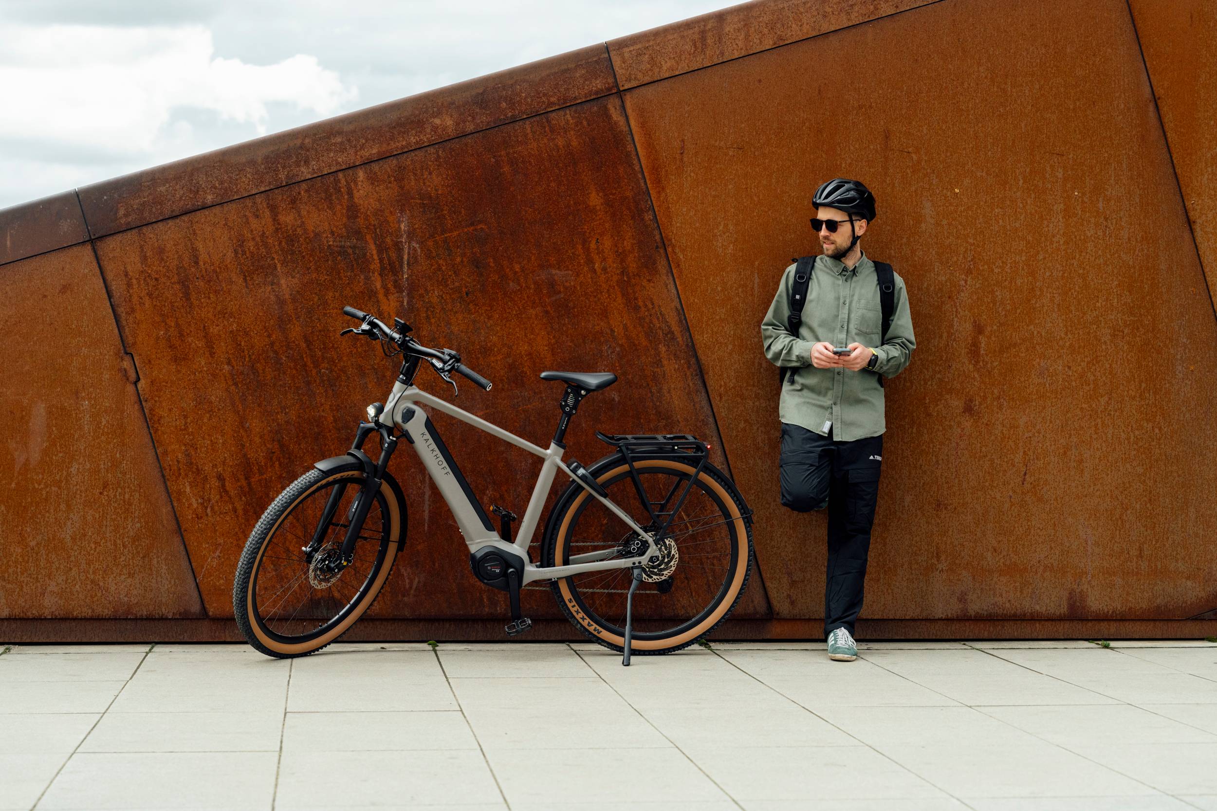 man with helmet and backpack leaning on wall and standing next to Kalkhoff bike