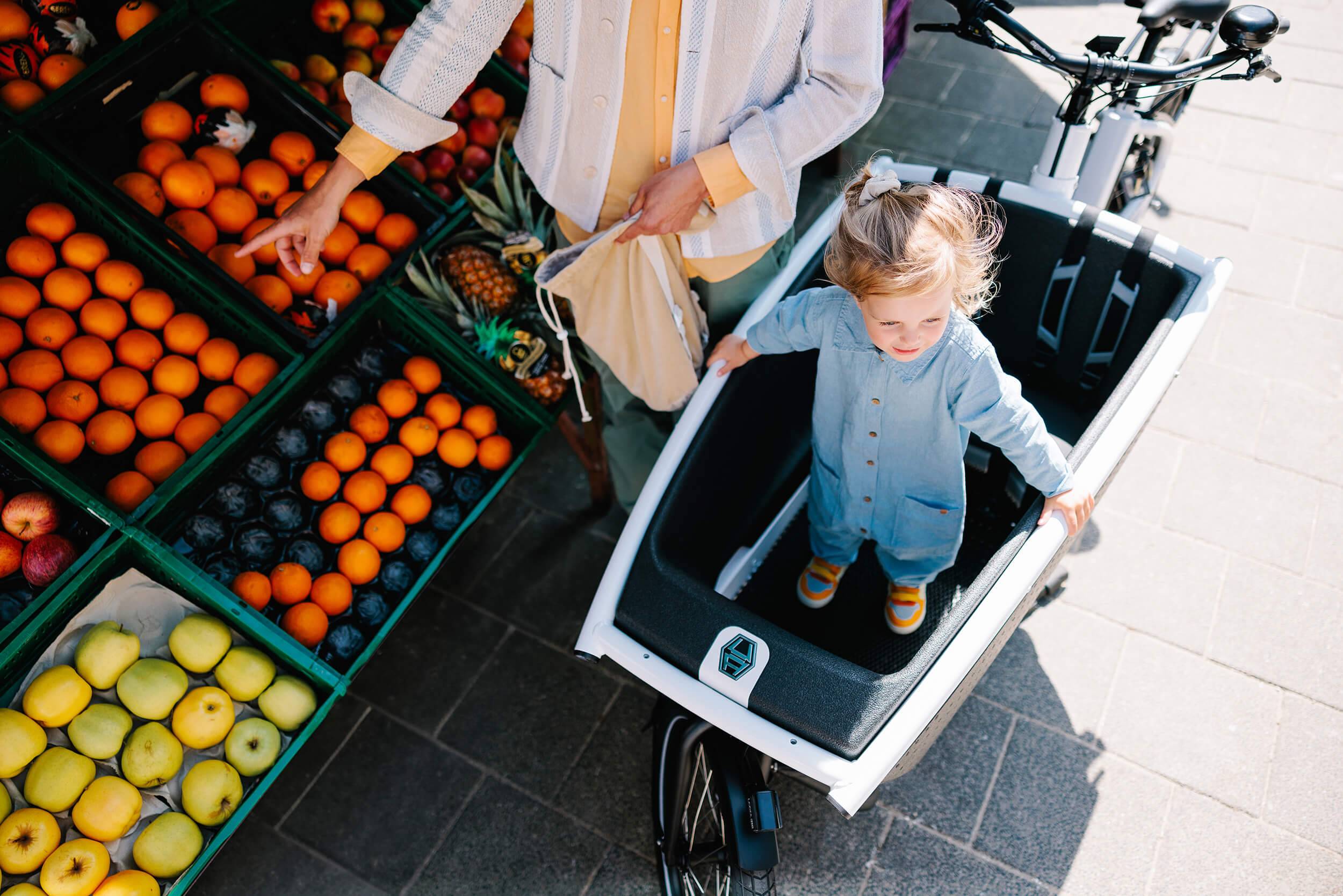 kid is standing in cargo bike while woman is buying fruits