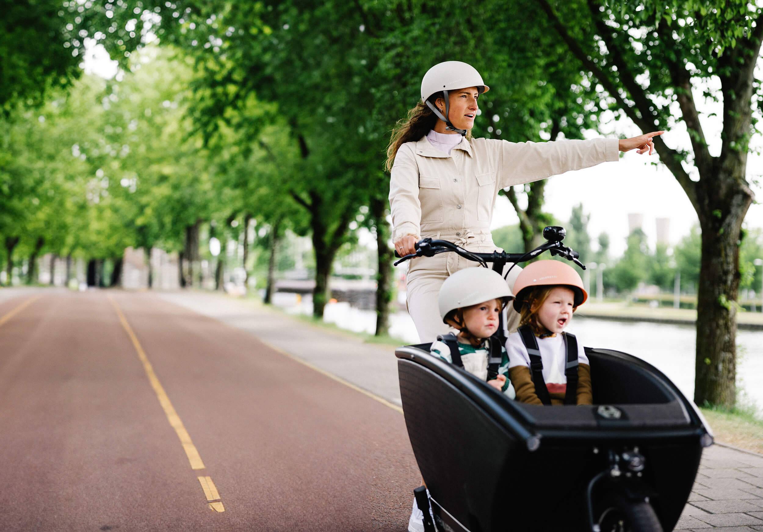 Frau mit zwei Kindern auf Lastenrad auf Radweg nahe Wasser zeigt auf etwas