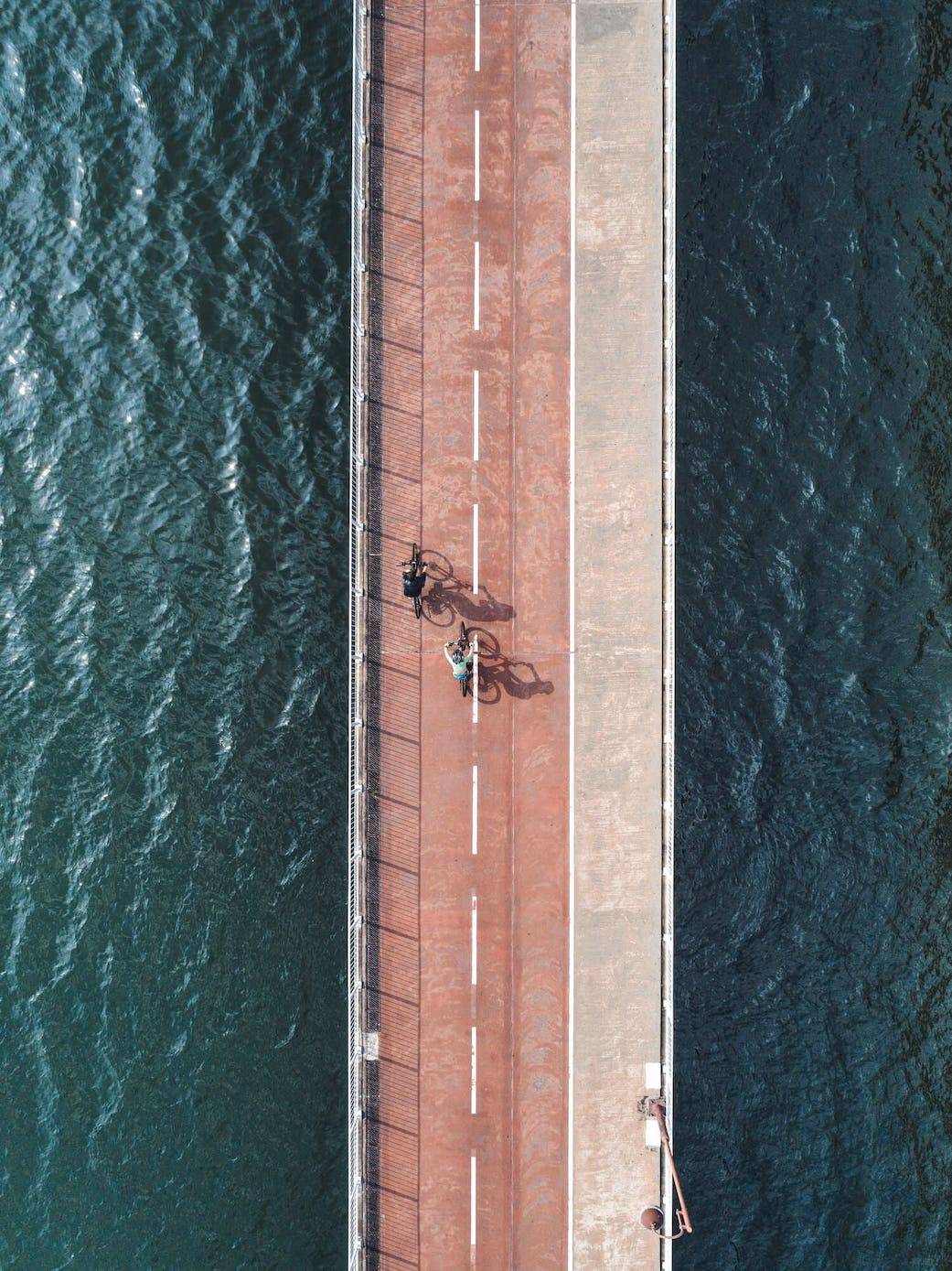 Two bikers riding their bikes on street with water on left and right side